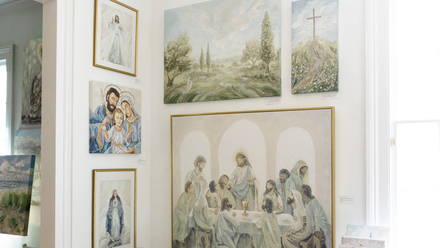 A styled interior room displaying a collection of religious art prints and paintings, including a large central print of the Last Supper, smaller prints of the Virgin Mary, Jesus, and a cross, arranged on white walls above a white decorative cart and wooden floor.